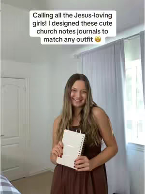 A young woman with long, light brown hair smiles while holding a white notebook in front of her. She's wearing a brown dress and standing in a room with white walls, a bed with a plaid blanket visible in the bottom left corner and a window with a white curtain on the right.