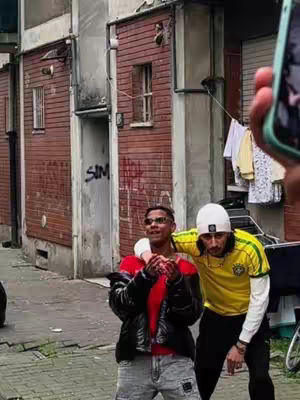 Two young men are standing on a street corner in front of a brick building. One is wearing sunglasses, a red shirt, and a black jacket. The other is wearing a white beanie and a yellow and green soccer jersey. The photo is taken seemingly from a phone.