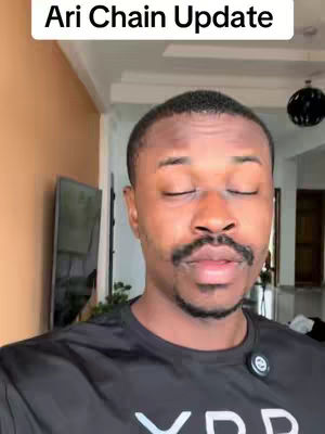 The image features a man with closed eyes, set against a neutral indoor background. He is wearing a black t-shirt with the word 'XRP' visible in white letters. The lighting suggests an interior setting. A white rectangular banner with the text 'Ari Chain Update' is located above the man's head.