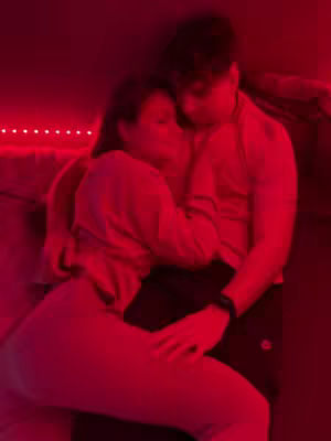A couple is embracing in a dim room lit with red light, the image is slightly blurred. They are tightly hugging, the woman is in the foreground, and the man is behind her. The lighting creates a warm, intimate atmosphere.