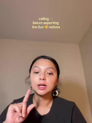 A woman with dark hair and a black top is in a selfie-style video. Text overlays read 'calling', 'Saturn aspecting', and 'the Sun natives'. She is gesturing with her index finger raised. The background is a plain wall.
