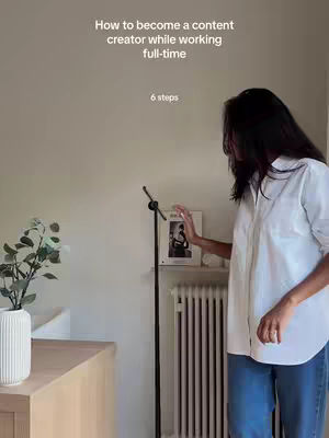 The cover image shows a woman standing in a well-lit indoor space. She is partially visible from her back, facing towards the left. She's wearing a white button-down shirt and blue jeans. The text overlay, placed at the top, reads: 'How to become a content creator while working full-time'. There's a plant in a white vase on a wooden side table featured on the left, with what appears to be a framed picture, a microphone and a radiator against the off-white wall. The overall composition is balanced and clean.