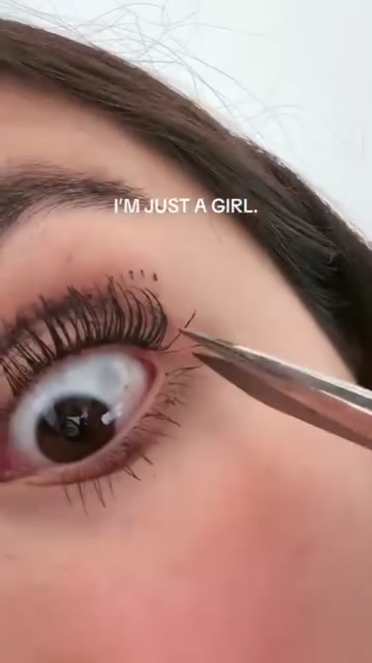 Close-up shot of an eye with dramatic makeup, focused on the eye’s detail. A pair of tweezers are held near the eye, potentially cutting a stray lash. Above the eye, text overlayed reads, “I’M JUST A GIRL.” Overall, it’s bright and shows the details of the eye.