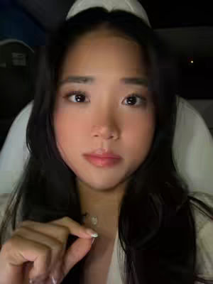 A close-up selfie of a young woman with long dark hair looking directly at the camera. She has a fair complexion, full lips, and is wearing subtle makeup. She's holding a small object in her fingers, and the background is softly out of focus.