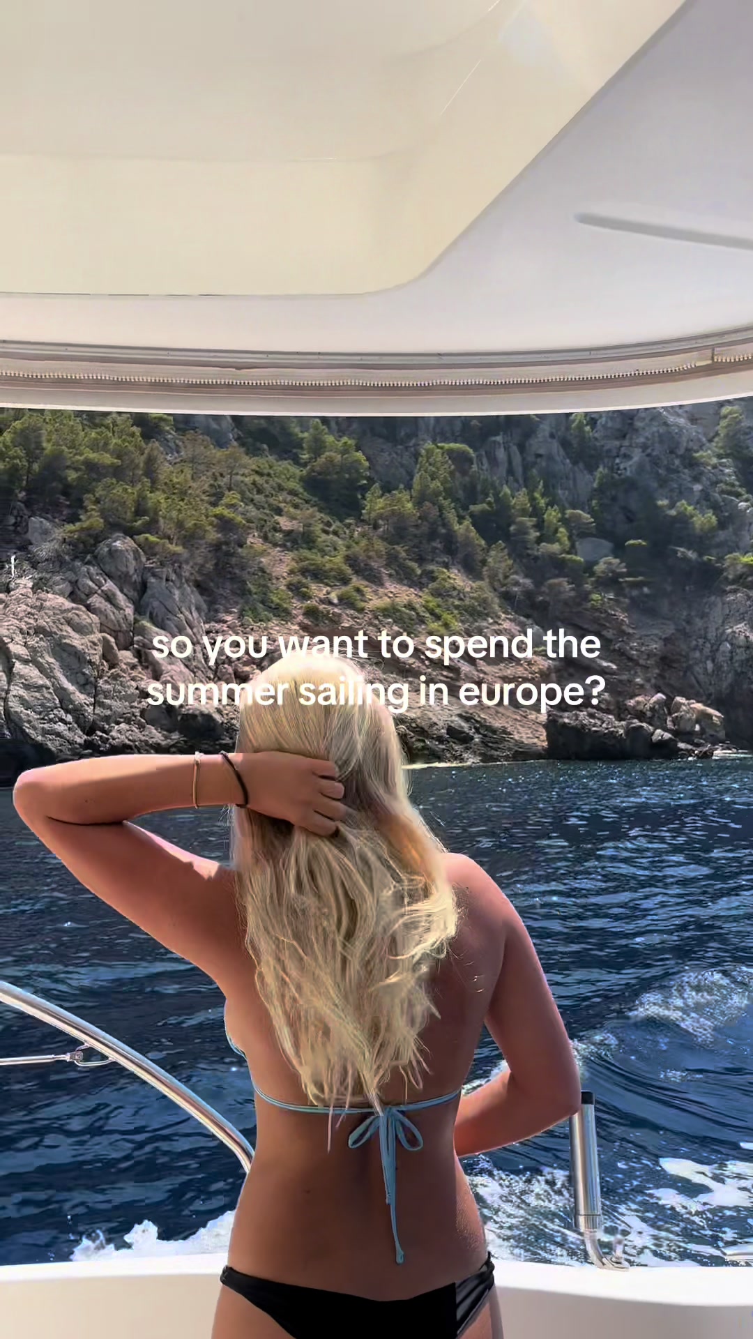 A woman with long blonde hair, wearing a blue bikini, stands on a boat looking out at a view of mountains and water. The boat has white railings and a white roof. The water is a deep blue, and the mountains are green and gray.
