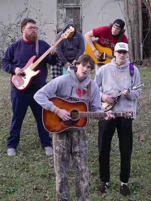 A group of five young men stand outdoors with guitars and an instrument, presumably a band, The central figure in the front holds an acoustic guitar and is wearing camo pants and grey hoodie, a bass guitar is in the second from left, an electric guitar in the second from right, and a white hat is worn by the one to the right with a mandolin in hand. The background shows a building and greenery.