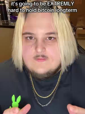 A close-up shot features a person with fair skin and light-blonde, chin-length hair. They wear a black t-shirt with a small green design, two necklaces and have a slight mustache and beard. The background is slightly blurred, suggesting an indoor setting.