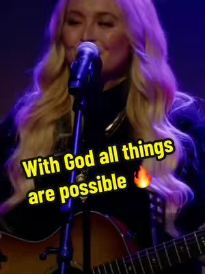 A woman with long blonde hair is singing into a microphone while holding an acoustic guitar. The background is dark, with purple lighting. The image has a text overlay with a motivational quote.