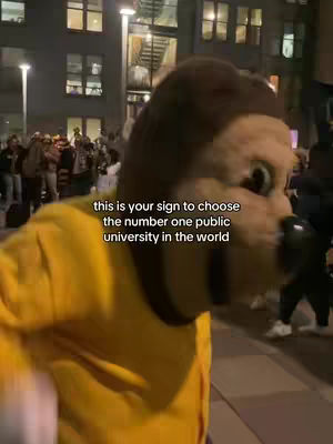 The cover image shows a person in a lion mascot head, standing in front of a building with a crowd of people in the background. The person is wearing a yellow shirt with a dark collar. The text overlay says: "this is your sign to choose the number one public university in the world".