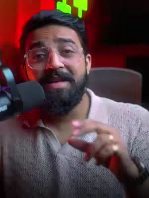 A man with glasses and a beard speaks into a microphone. The background has red and green lighting. The man is dressed in a light color shirt.