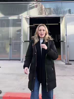 A woman stands in front of a building with glass doors and a metal frame, holding a microphone. The woman wears a black coat, white shirt, and blue jeans. The image has a bright, natural lighting, and the background is slightly blurred.