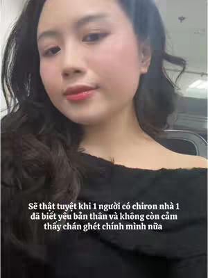 A selfie-style shot featuring a young woman with dark hair. She has fair skin and is wearing some makeup, including blush and red lipstick. The background is blurred out, but there appears to be some type of indoor setting. The lighting seems soft and natural. Text overlay is present at the bottom of the frame.