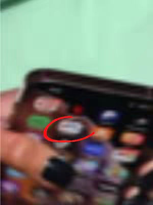 The image captures a smartphone screen displaying an app icon within a circular red highlight. The background is a soft, muted green. The phone is held in a hand, with the arm and a black watch visible, all slightly out of focus.
