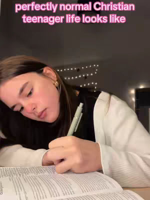 A young woman with long brown hair is sitting in a dimly lit room, presumably at a desk. She is leaning over an open book, with a pen in her right hand, appearing to be taking notes. The overhead text reads 'perfectly normal Christian teenager life looks like,' which is placed at the top. The background is blurry, but there are lights and other indistinct objects. The lighting is soft, with a focus on the woman's face and the book.