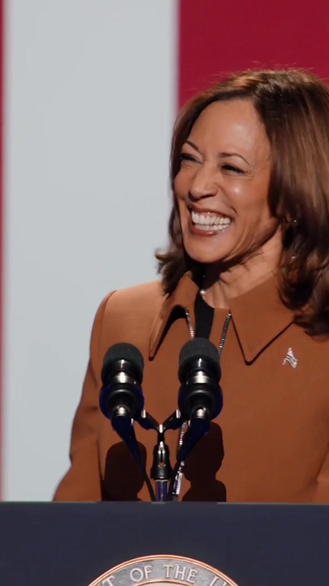 The image captures a medium shot of a woman, likely addressing an audience. She has brown hair and is wearing a brown blazer. The background features a white and red vertical panel. In the foreground, microphones are positioned, and the woman is smiling.