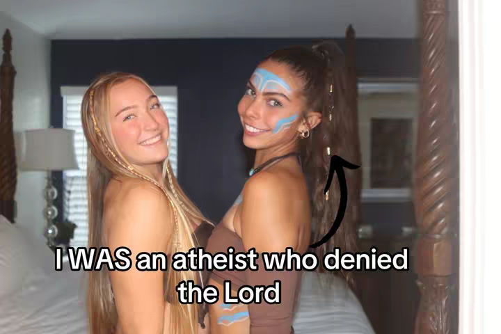 Two young women are smiling and posing together. One has blonde hair with braids, and the other has dark hair pulled back in a high ponytail with makeup, specifically light blue face paint. The women are standing indoors, with a bed and window visible in the background. The image is well-lit, and the composition focuses on the two individuals and their friendly interaction.