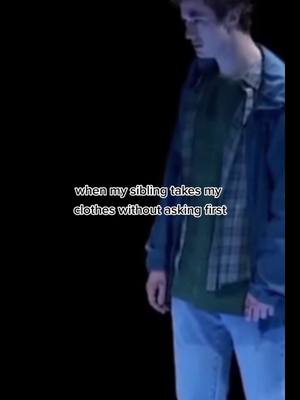 The image shows a young man in a casual outfit, standing slightly off-center against a dark, nearly black, background. The man is wearing a blue jacket over a green shirt and a plaid shirt. Text overlayed on the image reads 'when my sibling takes my clothes without asking first'. The lighting is dim, and the color palette is cool with dominant blues and greens.