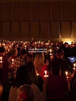 A low-angle shot captures a large gathering of people, seen from the back, holding lit candles. The lighting is dim, creating a warm, golden glow from the candles. The background shows a large space with wall panels. The text overlay reads, "never forget the true meaning of Christmas."