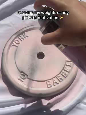 A close-up image shows a hand spraying a pink aerosol paint onto a weight plate. The weight plate is light gray in color with the words 'YORK BARBELL' imprinted on it. There's also text on the weight '10', implying its weight. The hand holding the spray can is the main point of focus and part is blurred; the background is a wrinkled white surface.