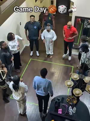 An overhead shot shows a family gathered indoors, likely for a game. A green box is drawn on the floor, possibly for a game, with people surrounding it. The image is well-lit, and the setting appears to be a home.
