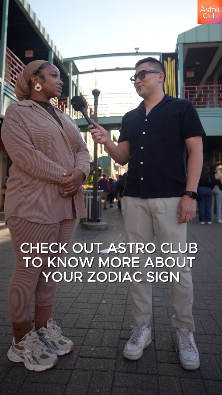A street scene featuring a person interviewing a woman in front of a blurred background. The woman, wearing a headscarf and beige outfit, speaks into the mic. The interviewer is wearing glasses and a dark shirt. Text overlay advertises an astrology app.