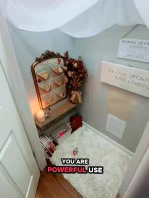 An interior shot of a small alcove or prayer space. The space is painted in light cool tones. A small shelf holds a candle, perfume bottles, and books. Above, a large mirror with a gold ornate frame, holding index cards. A white fluffy rug covers the floor. Text overlayed on the image.