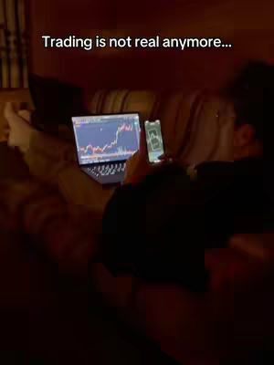 A person lies on a couch, using a laptop and a phone to display trading charts. The image is dimly lit, suggesting a relaxed setting. A text overlay states, "Trading is not real anymore..."