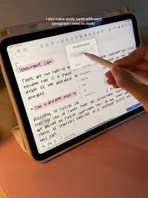 A person's hand holding an Apple Pencil is interacting with an iPad. The iPad is displaying notes about "Inheritance Law" with handwritten annotations and highlighted sections. The background includes a pink desk surface and a neutral-colored case for the iPad. The image composition is angled to focus on the notes and the user's interaction with the screen.