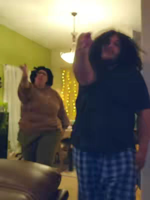 Two people are in a room; one is close to the camera and in the foreground, with their arm raised. The other, further back, also has their arm raised. The room is lit with a overhead light, and fairy lights in the background. The walls are painted in a cool tone and appear to be a residential setting.