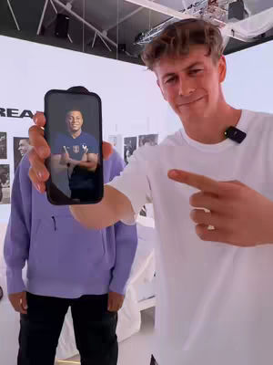 A man in a white t-shirt holds a phone displaying a picture of another man in a blue shirt. He points with his right index finger to the phone. Another person is seen in the background, wearing a purple hoodie.