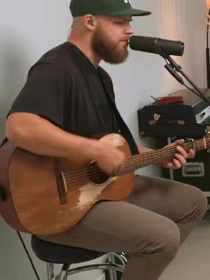 A man with a beard is seated, playing a worn acoustic guitar. He wears a black t-shirt, brown pants, and a green cap. The background is a neutral white wash.