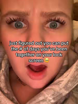 A close-up shot of a young woman's face, capturing her wide-eyed expression of surprise or excitement. Her eyes are the focal point, with defined lashes and a slightly open mouth. Overlaid on the image is text in a clean, modern font, indicating that she has discovered a way to display the number of days she's been together with someone on her lock screen. The overall composition is simple, with the woman's skin tone being the dominant color, along with the text.