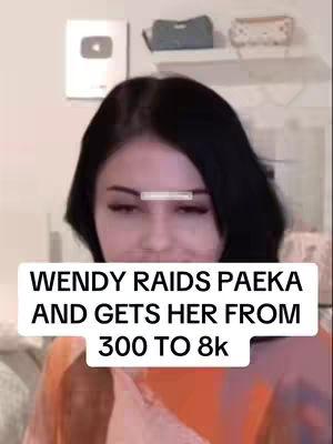 A selfie-style video cover features a woman with dark hair, visible makeup, and an orange top, filling most of the frame. A text overlay in a white box displays the title. Background elements include a shelf and a YouTube play button, suggesting a vlogging or social media context.
