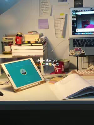 A well-lit desk setup with a laptop, tablet, books, and stationery. A tablet is at the bottom left, a laptop is on the top right, and there are books and other items adding visual interest.