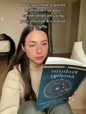 A young woman is seated indoors, holding a book titled "Predictive Astrology" with a dark blue cover. The background and her attire are of neutral tones, and the text overlay reads, "POV: you have a pisces midheaven so your career relies on using your intuitive & creative gifts." She looks directly at the camera, with a serious expression.