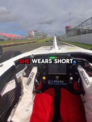 The cover image shows a first-person view from inside a race car on a track. The steering wheel and the driver's gloved hands are in focus, the red seat harness is visible, and parts of the car's interior and dashboard are in view. The track curves away in the distance and a sky with clouds can be seen. Text layers are added to the image.