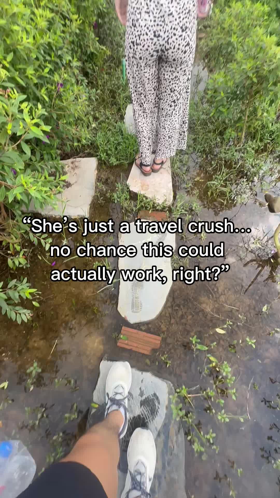 The cover image is a first-person perspective shot of someone walking on stepping stones across a shallow stream or pond. Overhanging vegetation and greenery surround the water and the walkway. In the foreground, the viewer sees their own feet in white sneakers, while in the mid-ground, the legs of someone else are shown wearing patterned pants. The background shows more foliage and a small view of the sky.