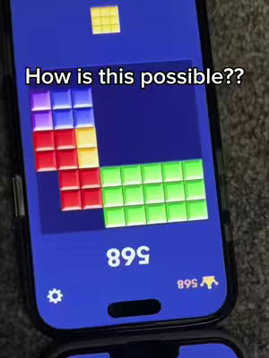 A phone screen displays a puzzle game with blocks of colored squares in progress. The phone is held at an angle, and the background is a blurry surface. There is also a text overlay.