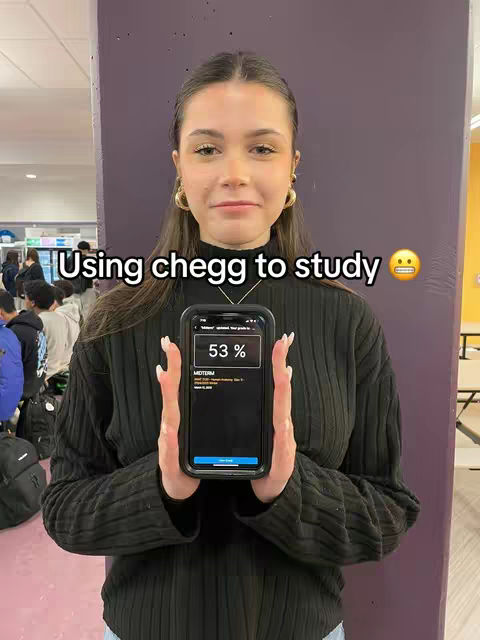 A young woman with fair skin and long hair holds a smartphone in front of a purple background. The phone displays a screen with the words and a percentage. Text overlays above the woman that says, "Using chegg to study". The woman wears long-sleeve black tops and a silver necklace. She stands in front of the camera.