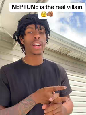 A young Black man with short dreadlocks is the central focus, set against a casual outdoor background. He wears a black t-shirt, a gold chain, and a black head wrap. The overlay text states 'NEPTUNE is the real villain' in white, positioned above his head. The background consists of a light-colored wall and a partly visible sky. The man's facial expression and hand gestures suggest he is speaking, and the overall layout focuses on delivering a direct message.