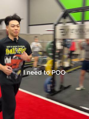 A man running towards the camera with a gym environment in the background. The man is wearing a black graphic tee and black pants. The gym has exercise equipment and a red floor.
