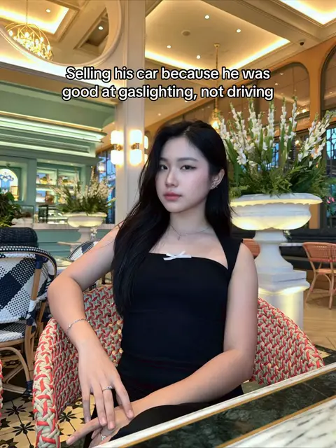 A woman sits in a chair, looking directly at the camera. She is wearing a black, square-neck top and has long, dark hair. The background is a restaurant or similar setting, with decorations and natural light.
