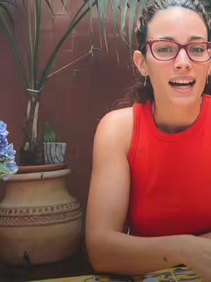 The cover image features a woman with dark curly hair, wearing a red tank top and glasses. She is positioned in front of a blurred outdoor background with a potted plant to her left. She is smiling and looking towards the camera.