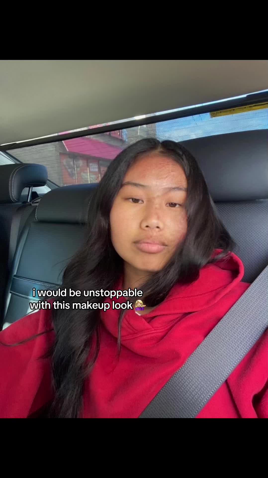 The cover image shows a young woman inside a car. She's wearing a red hoodie and facing the camera directly. The background is blurred, suggesting movement and includes a glimpse of an urban setting. The lighting appears to be natural, and the composition is centered on the person. Text overlay stating 'i would be unstoppable with this makeup look' is present on the screen.