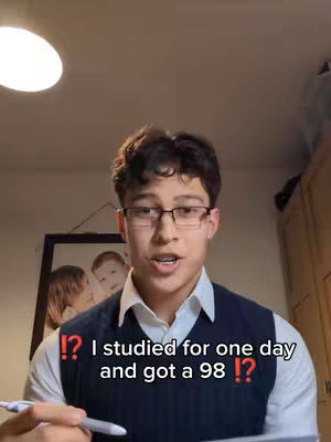 A young man with glasses is talking to the camera. The background includes a light, a framed picture of children, and part of a cabinet. Text added to the image states, "I studied for one day and got a 98!?"