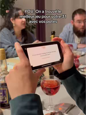 A person holds a phone displaying a game question in front of a group of people at a table, with a glass of red-colored drink in the foreground.