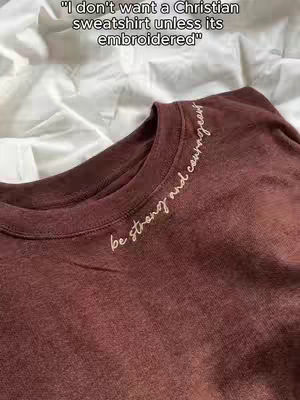 The cover image features a close-up of a maroon sweatshirt laid on top of a white, crinkled surface. White text is embroidered along the neckline of the sweatshirt, reading "be strong and courageous". Above the sweatshirt, in a sans-serif font, is the text "I don't want a Christian sweatshirt unless its embroidered"
