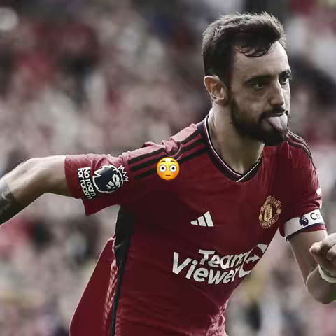 The image captures a right-side profile of a soccer player in action. He's wearing a red jersey with black stripes on the shoulders, and the Manchester United crest is visible. The player has short, dark hair and has his tongue out slightly. An emoji is placed next to his shoulder.