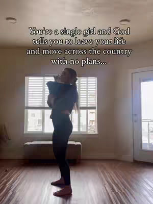 A woman stands in a room with large windows and holds something in her arms, possibly a pet or small child. The room has a neutral color palette with wooden floors, a beige bench under the window, and a simple door. The main subject is centered against the backdrop of the window.