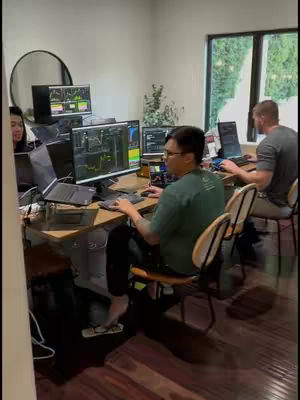 A group of people in a modern office setting, presumably day traders, are gathered around multiple computer screens displaying financial charts and graphs. The room has a minimalist aesthetic with natural light coming from a large window. The main subject, a man wearing glasses and a green shirt, is focused on a computer, and multiple other people are working on laptops.